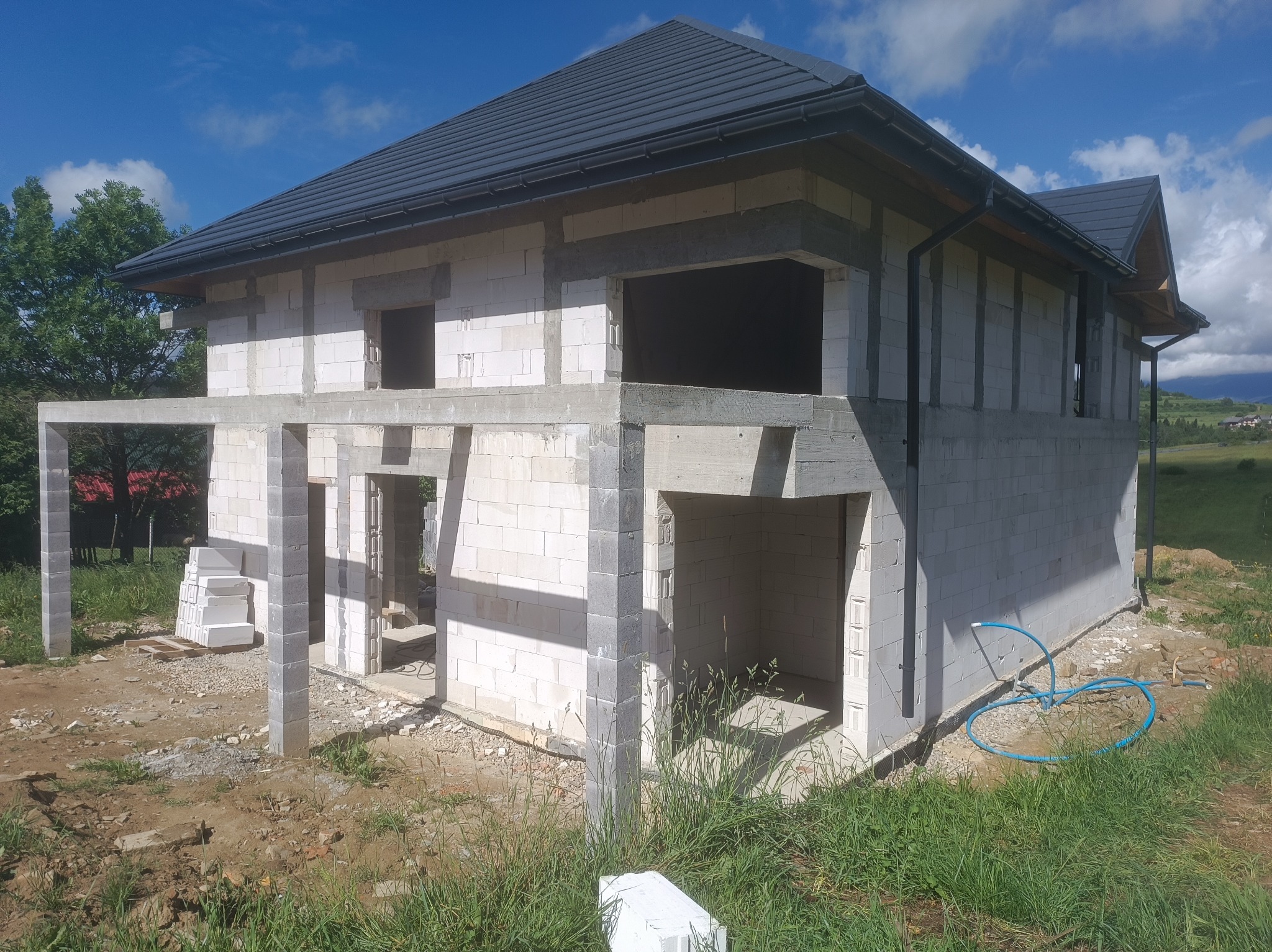 Surowy stan domu z betonu komórkowego pod ciemnym dachem, widoczne otwory okienne i drzwiowe, teren wokół w stanie budowy, błękitne niebo w tle.