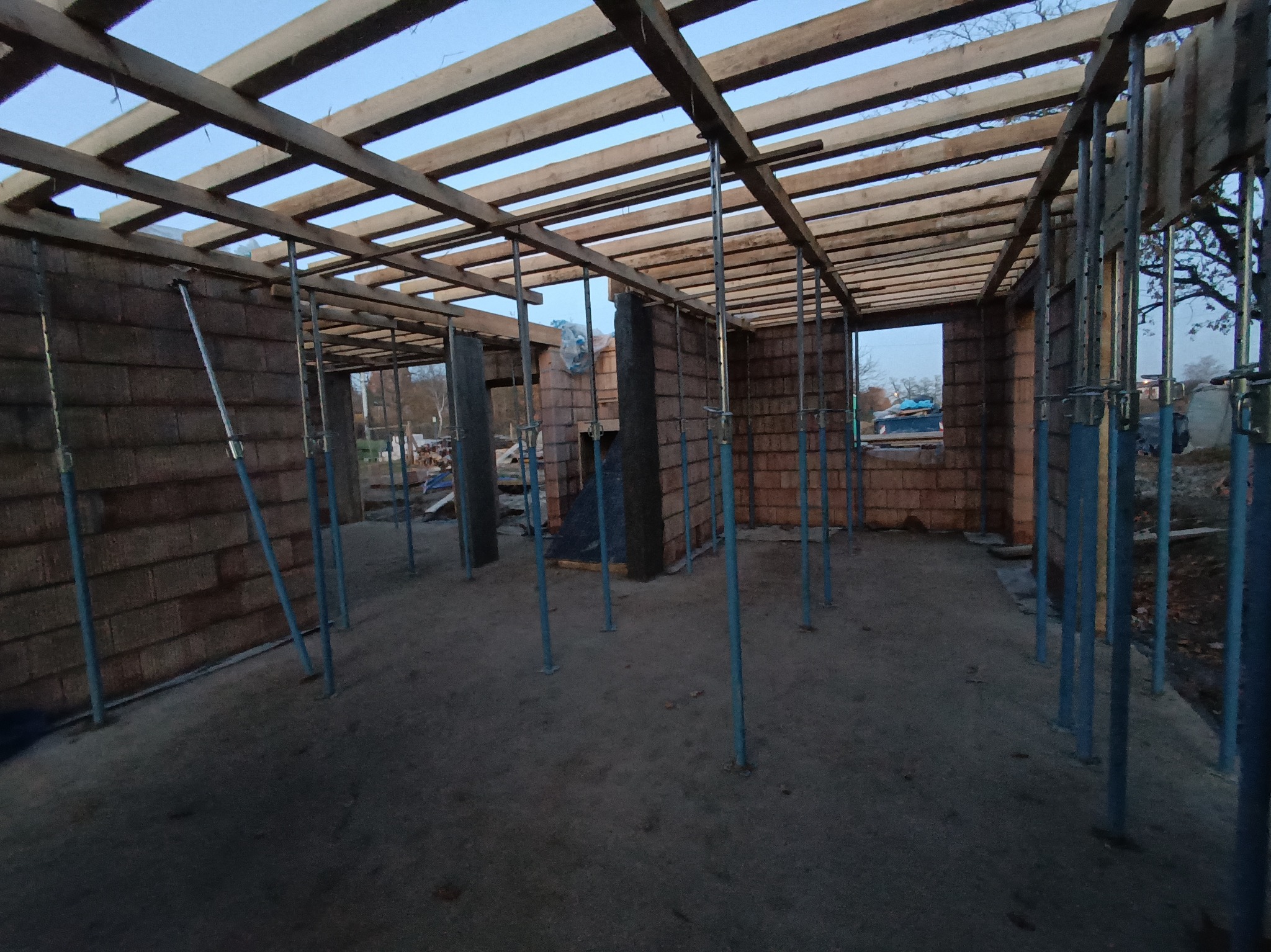 Wnętrze budynku w trakcie budowy z cegieł, z drewnianą konstrukcją dachu i niebieskimi podporami. Widoczne otoczenie zewnętrzne przez otwory okienne.