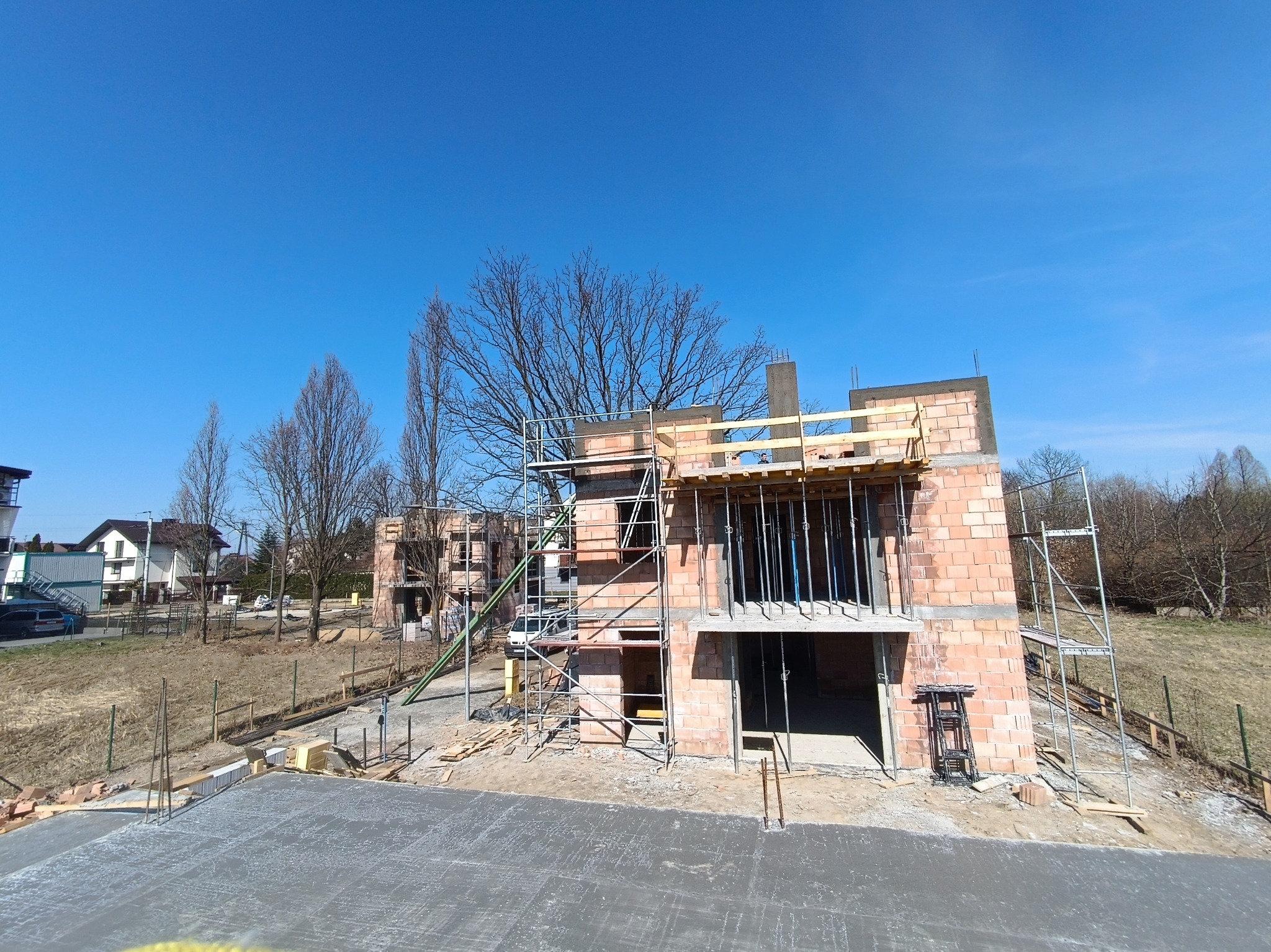 Budowa domu z czerwonej cegły w słoneczny dzień. Widoczne rusztowania, betonowa podstawa i elementy konstrukcyjne. W tle drzewa i błękitne niebo.