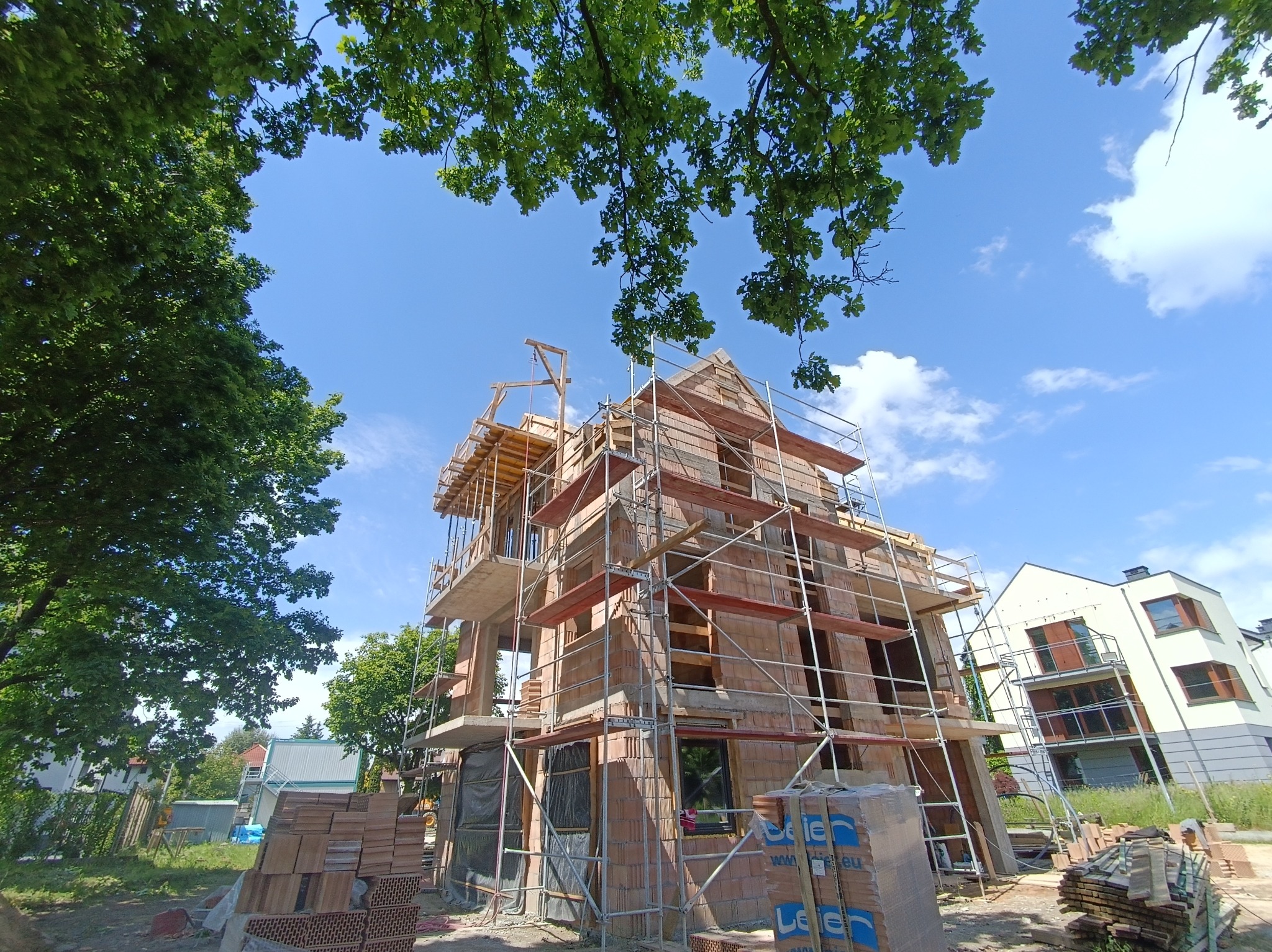 Wznoszenie domu z cegły, widok z dołu na rusztowania i fragmenty zieleni. Niebo błękitne, częściowo zasłonięte liśćmi drzew. Cegły ułożone w paletach na pierwszym planie.