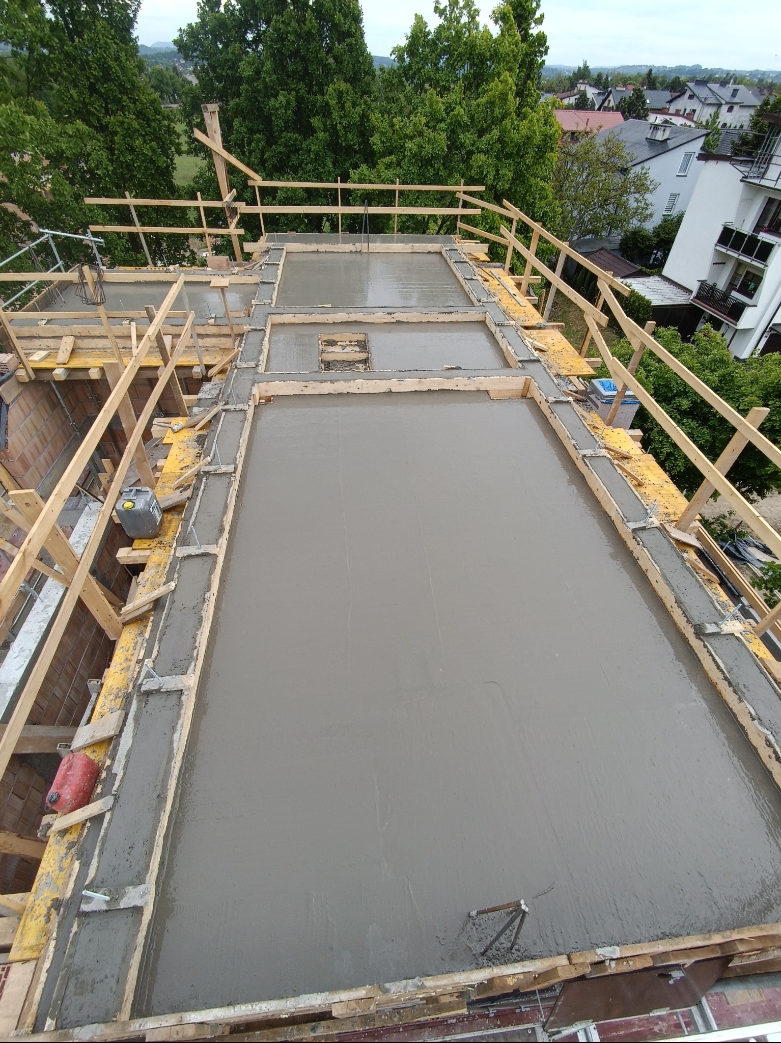 Zalany beton na stropie budynku w trakcie budowy, widok z góry. Szalunki drewniane, częściowo widoczne budynki w tle. Świeży beton na całej powierzchni.