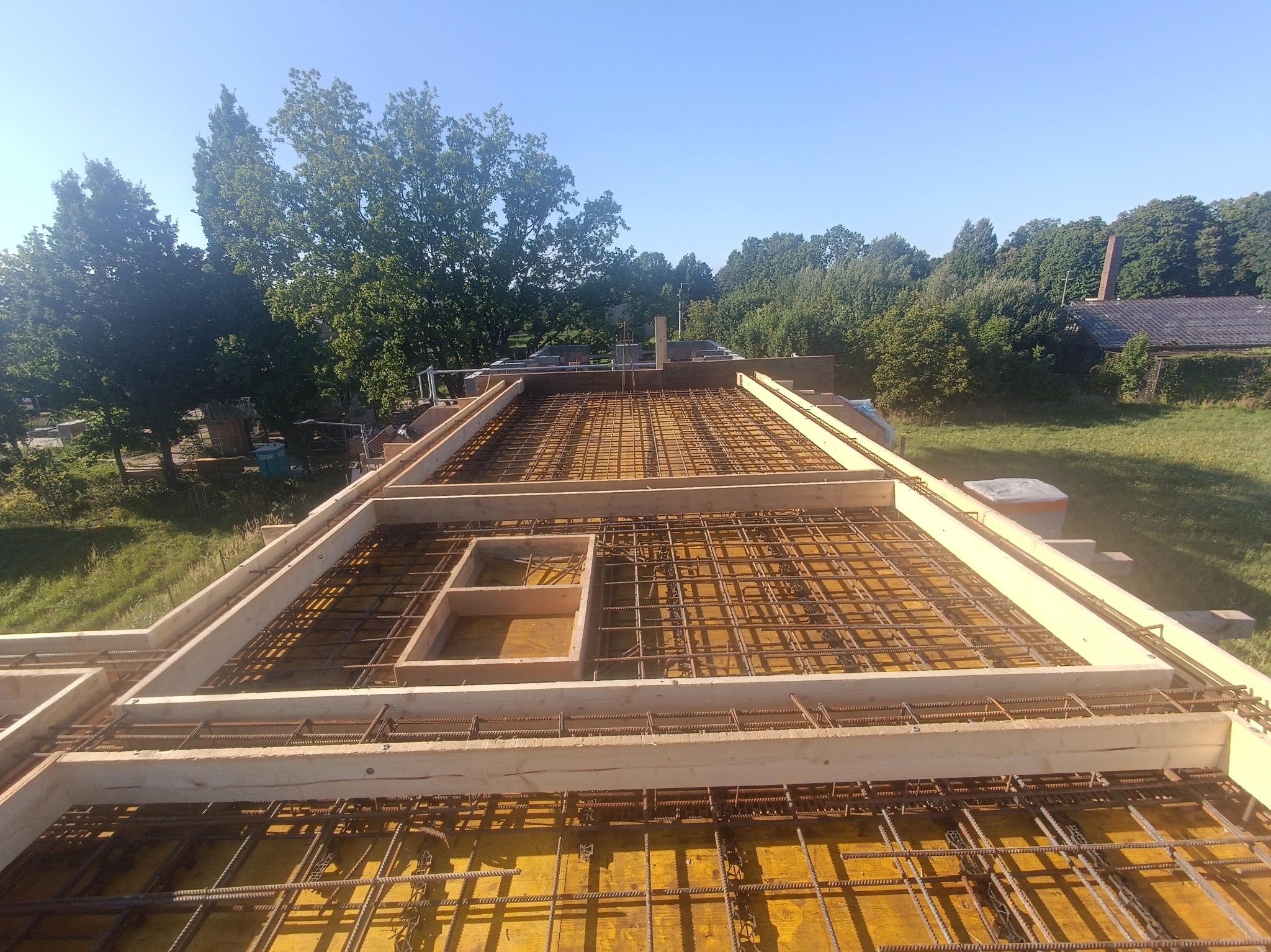 Zbrojenie stropu z drewnianym szalunkiem, widok z góry na siatkę z prętów stalowych i deski, w tle zieleń drzew i błękitne niebo. Etap budowy domu.