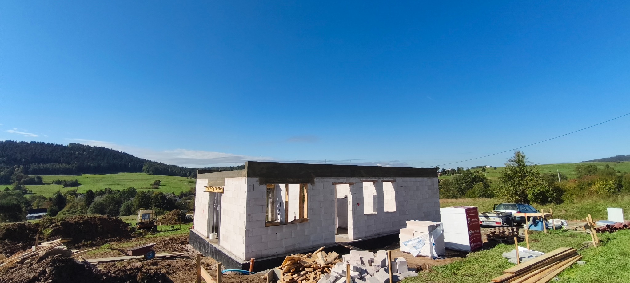 Budowa domu z betonu komórkowego w malowniczej scenerii. Widoczne ściany w stanie surowym otwarte, materiały budowlane i zielone wzgórza w tle.
