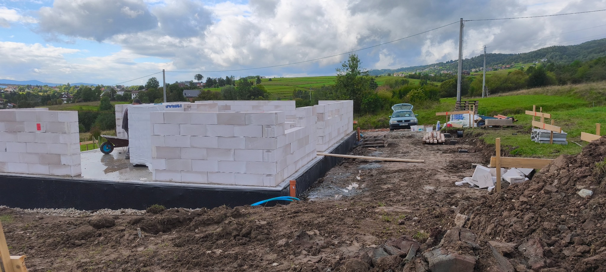 Budowa domu w toku: wzniesione ściany z białych bloczków na fundamencie z hydroizolacją. W tle krajobraz z zielonymi wzgórzami i częściowo widoczną zabudową.
