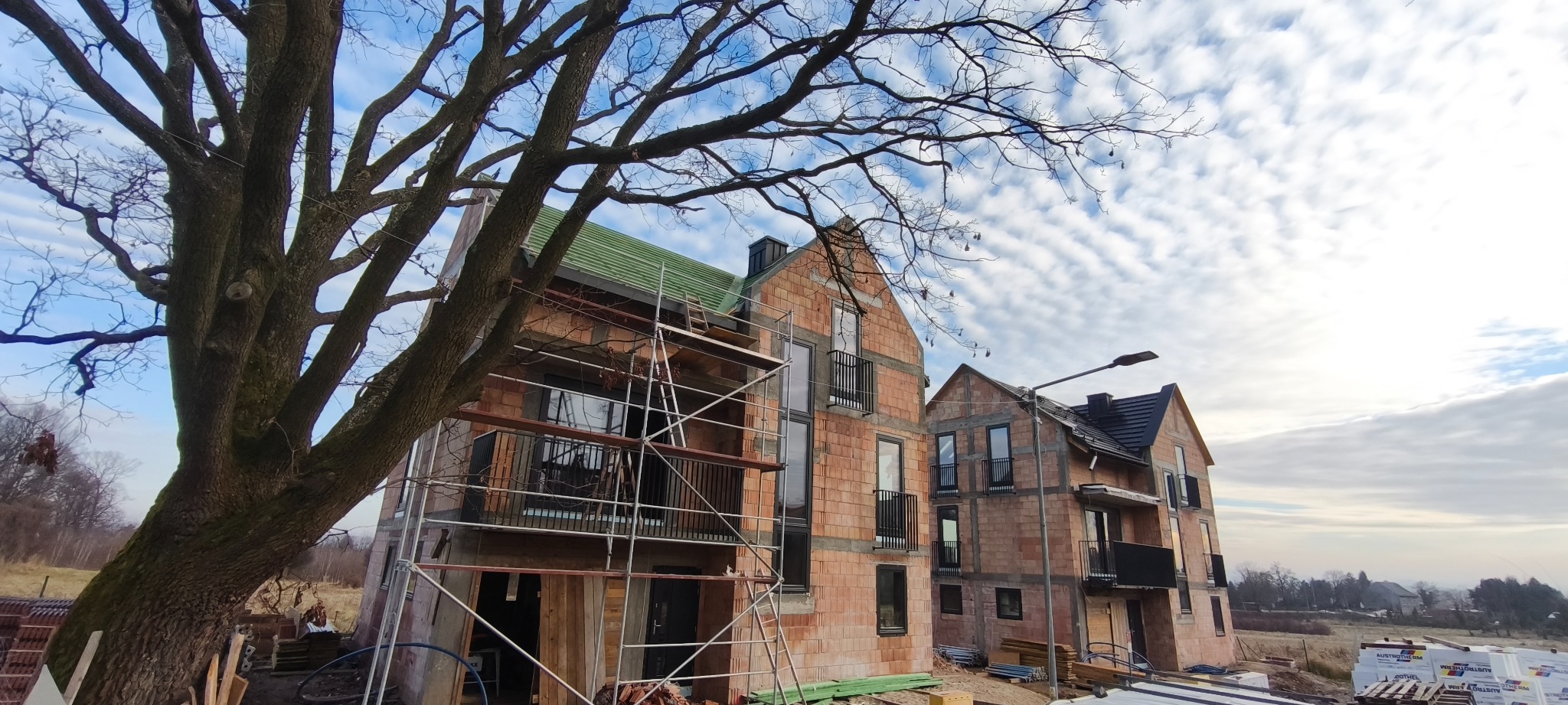 Dwa budynki w trakcie budowy z czerwonej cegły, z zielonym dachem na jednym i czarnym na drugim, otoczone rusztowaniami i drzewem bez liści na tle pochmurnego nieba.