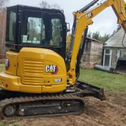 Żółta mini koparka CAT 303E na placu budowy, łyżka wypełniona ziemią, w tle budynek gospodarczy i drzewa.