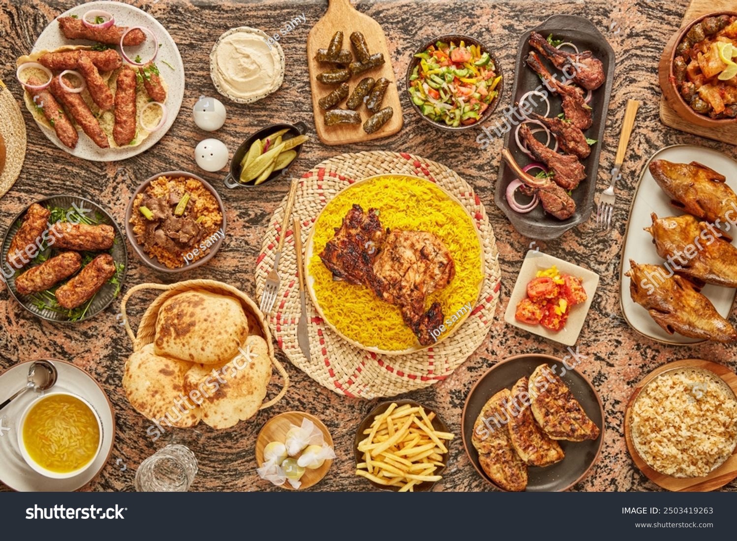 Bogato zastawiony stół z różnorodnymi daniami kuchni bliskowschodniej: grillowane mięsa, ryż, sałatki, pieczywo, frytki i przystawki, ułożone na marmurowym blacie i plecionych podkładkach.