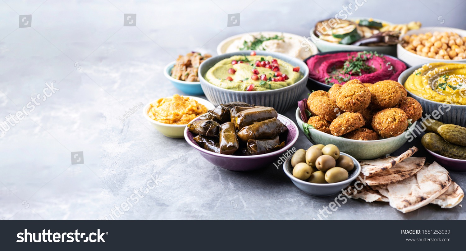 Bogaty wybór przystawek w miseczkach na szarym tle: hummus w różnych wariantach kolorystycznych, faszerowane liście winogron, falafel, oliwki, marynowane ogórki i chleb pita.