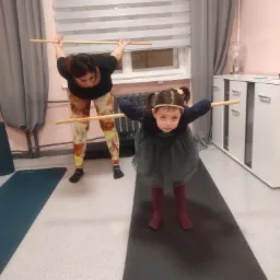 Kobieta i dziewczynka wykonują ćwiczenie z drewnianym kijem na macie do ćwiczeń w pomieszczeniu z roletami i komodą.