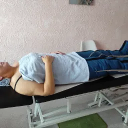 Kobieta leży na stole do masażu w kombinezonie do presoterapii, przykryta ręcznikiem, z ręcznikiem pod głową, w tle ściana z fakturą i fragment obrazu.