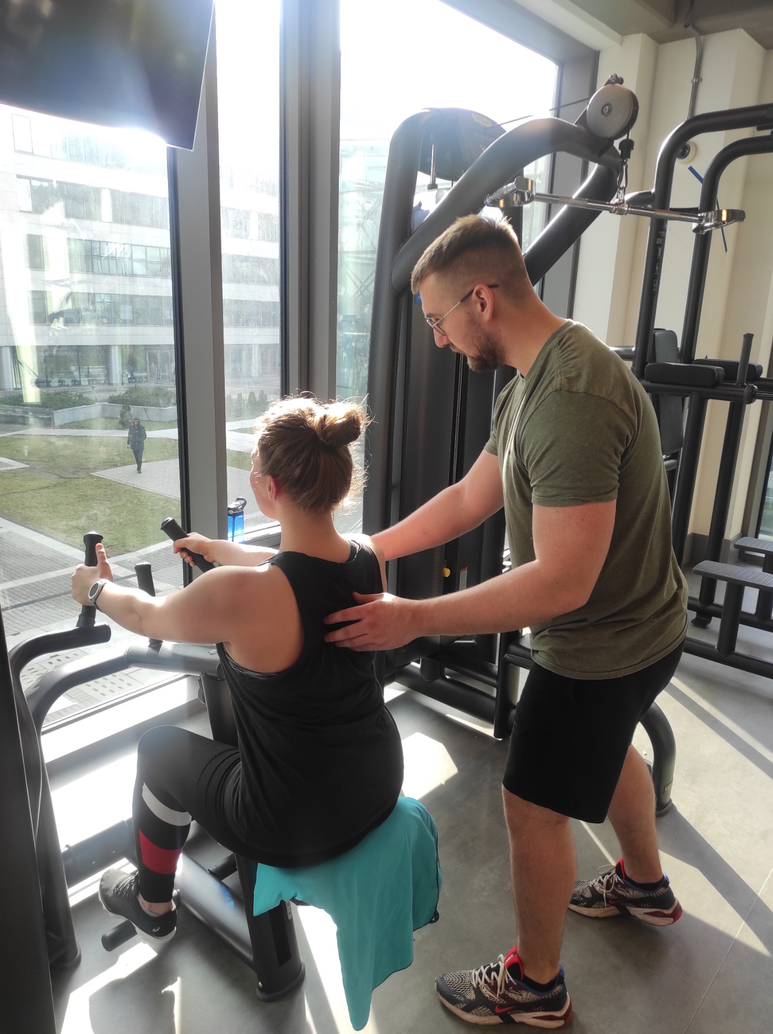 Kobieta ćwiczy na maszynie pod okiem trenera personalnego w jasnej siłowni z widokiem na park.