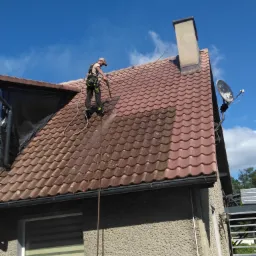 Pracownik w uprzęży, z dyszą ciśnieniową, czyści dach z czerwonej dachówki, widoczny kontrast czystej i zabrudzonej powierzchni, antena satelitarna i komin w tle.