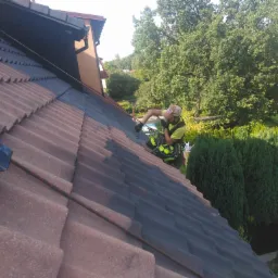 Pracownik w uprzęży, zabezpieczony linami, maluje fragment dachu z czerwonej dach&oacute;wki na czarno, widok z g&oacute;ry, w tle zieleń drzew i zabudowania.