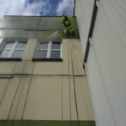 Pracownik w jaskrawozielonym kombinezonie, zawieszony na linach, spryskuje elewację budynku wodą pod ciśnieniem, czyszcząc ją od góry do dołu, widoczne okna i fragment dachu.