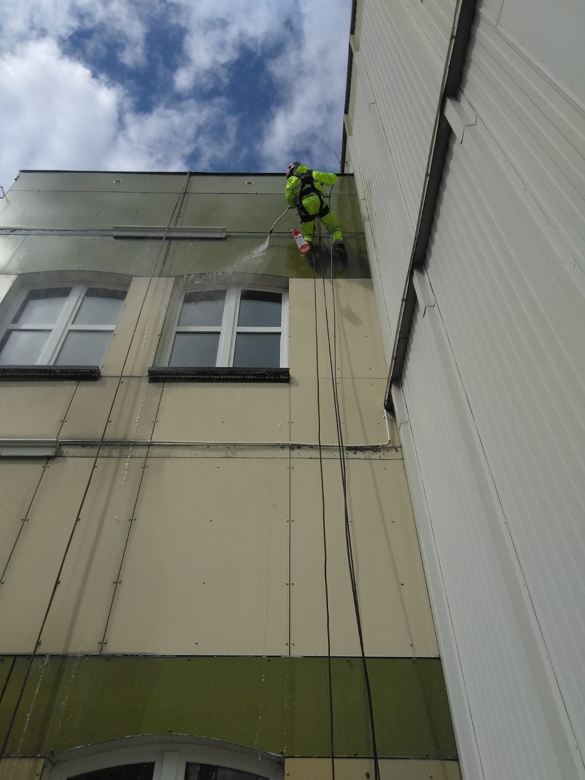 Pracownik w jaskrawozielonym kombinezonie, zawieszony na linach, spryskuje elewację budynku wodą pod ciśnieniem, czyszcząc ją od góry do dołu, widoczne okna i fragment dachu.
