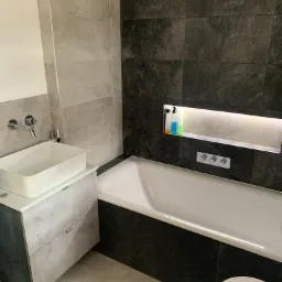 Nowoczesna łazienka z białą, prostokątną umywalką nablatową, szarą szafką pod umywalką, wanną obudowaną czarnymi płytkami, wnęką z podświetleniem i armaturą w kolorze chromu.