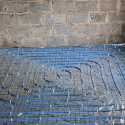 Niebieskie rurki ogrzewania podłogowego ułożone spiralnie na folii izolacyjnej z nadrukiem, przymocowane białymi opaskami do metalowej siatki na tle surowych ścian z bloczków betonowych.