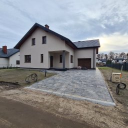 Bruk Montaż Układanie i Sprzedaż kostki brukowej Biała Podlaska - Nowoczesny dom jednorodzinny z szarą kostką brukową na podjeździe, widoczna skrzynka gazowa i fragmenty ogrodzenia. Pochmurne niebo w tle.
