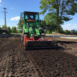 Bruk Montaż Układanie i Sprzedaż kostki brukowej Biała Podlaska - Zielony traktor z glebogryzarką spulchnia ciemną ziemię na działce. Operator w kamizelce odblaskowej. Błękitne niebo i drzewo w tle. Przygotowanie terenu pod uprawę.