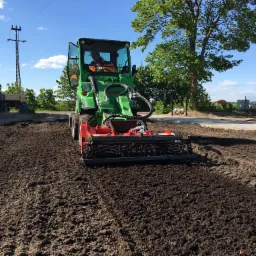 Zielony traktor z glebogryzarką spulchnia ciemną ziemię na działce. Operator w kamizelce odblaskowej. Błękitne niebo i drzewo w tle. Przygotowanie terenu pod uprawę.