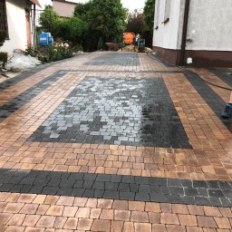 Bruk Montaż Układanie i Sprzedaż kostki brukowej Biała Podlaska - Wzorzysta nawierzchnia z kostki brukowej w odcieniach brązu i szarości, czyszczona wodą pod ciśnieniem. Widoczny fragment domu i zieleni w tle.