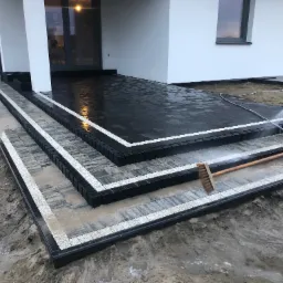 Nowoczesne schody wejściowe z kostki brukowej w odcieniach szarości i czerni, wykończone białym obramowaniem. Osoba czyści powierzchnię myjką ciśnieniową i szczotką.