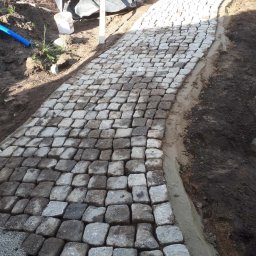 Bruk Montaż Układanie i Sprzedaż kostki brukowej Biała Podlaska - Ścieżka z kostki granitowej w trakcie układania, widoczne worki z materiałami budowlanymi w tle. Krawężnik z betonu. Częściowo ułożona nawierzchnia.
