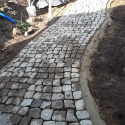 Ścieżka z kostki granitowej w trakcie układania, widoczne worki z materiałami budowlanymi w tle. Krawężnik z betonu. Częściowo ułożona nawierzchnia.