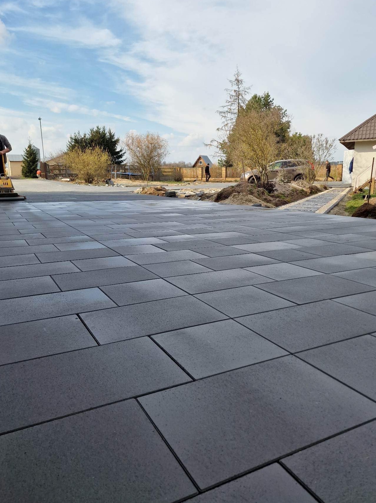 Ułożona, szara kostka brukowa na posesji. W tle widoczny dom, drzewa i częściowo ogrodzenie. Na kostce widoczne ślady wilgoci. Prace w toku, widoczna ubijarka.