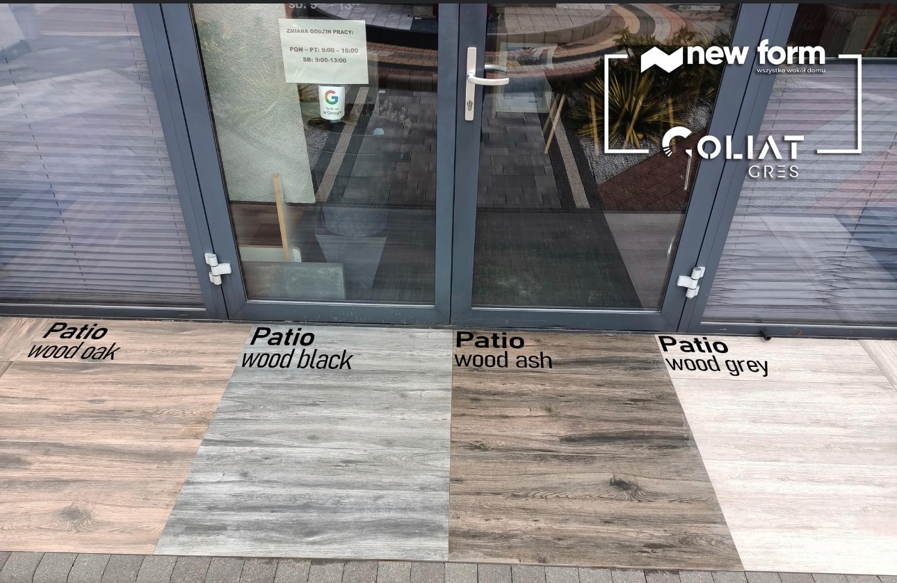 Cztery próbki płytek gresowych imitujących drewno (oak, black, ash, grey) ułożone przed wejściem z logo producenta 'New Form' i 'Goliath Gres' na szybie.