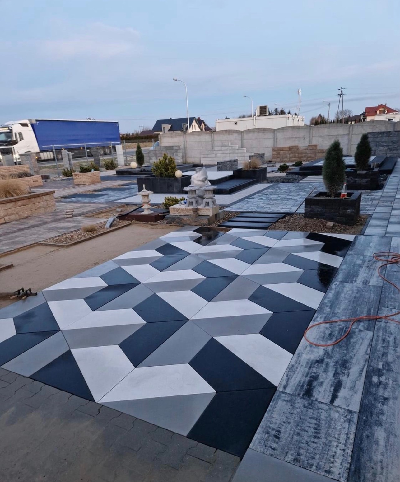 Efektowna, geometryczna kompozycja z kostki brukowej w odcieniach szarości i bieli, tworząca iluzję trójwymiarowości. Elementy małej architektury ogrodowej w tle.