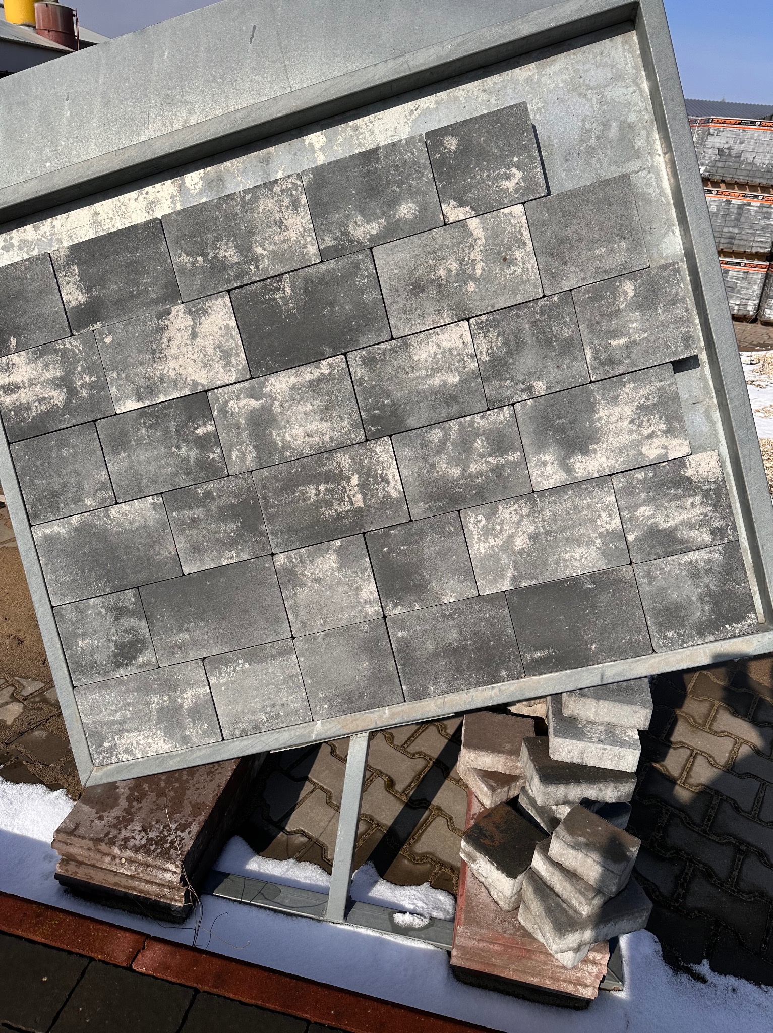 Prezentacja kostki brukowej w metalowej ramie, ukazująca wzór i fakturę. W tle widoczne palety z kostką oraz fragment zimowego krajobrazu.