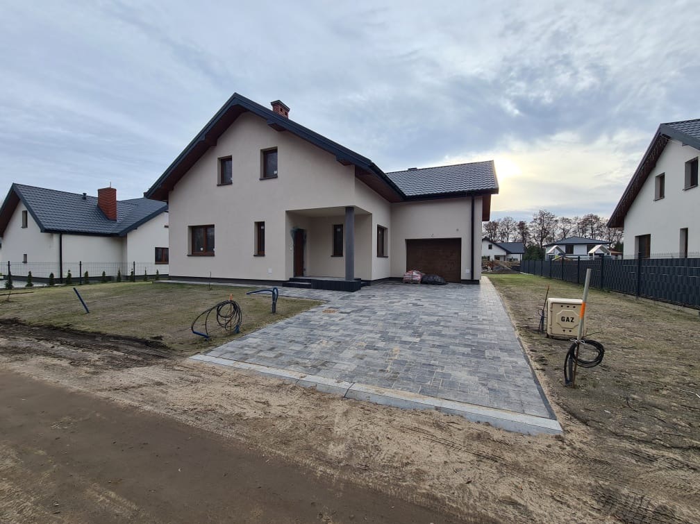 Nowoczesny dom jednorodzinny z szarą kostką brukową na podjeździe, widoczna skrzynka gazowa i fragmenty ogrodzenia. Pochmurne niebo w tle.