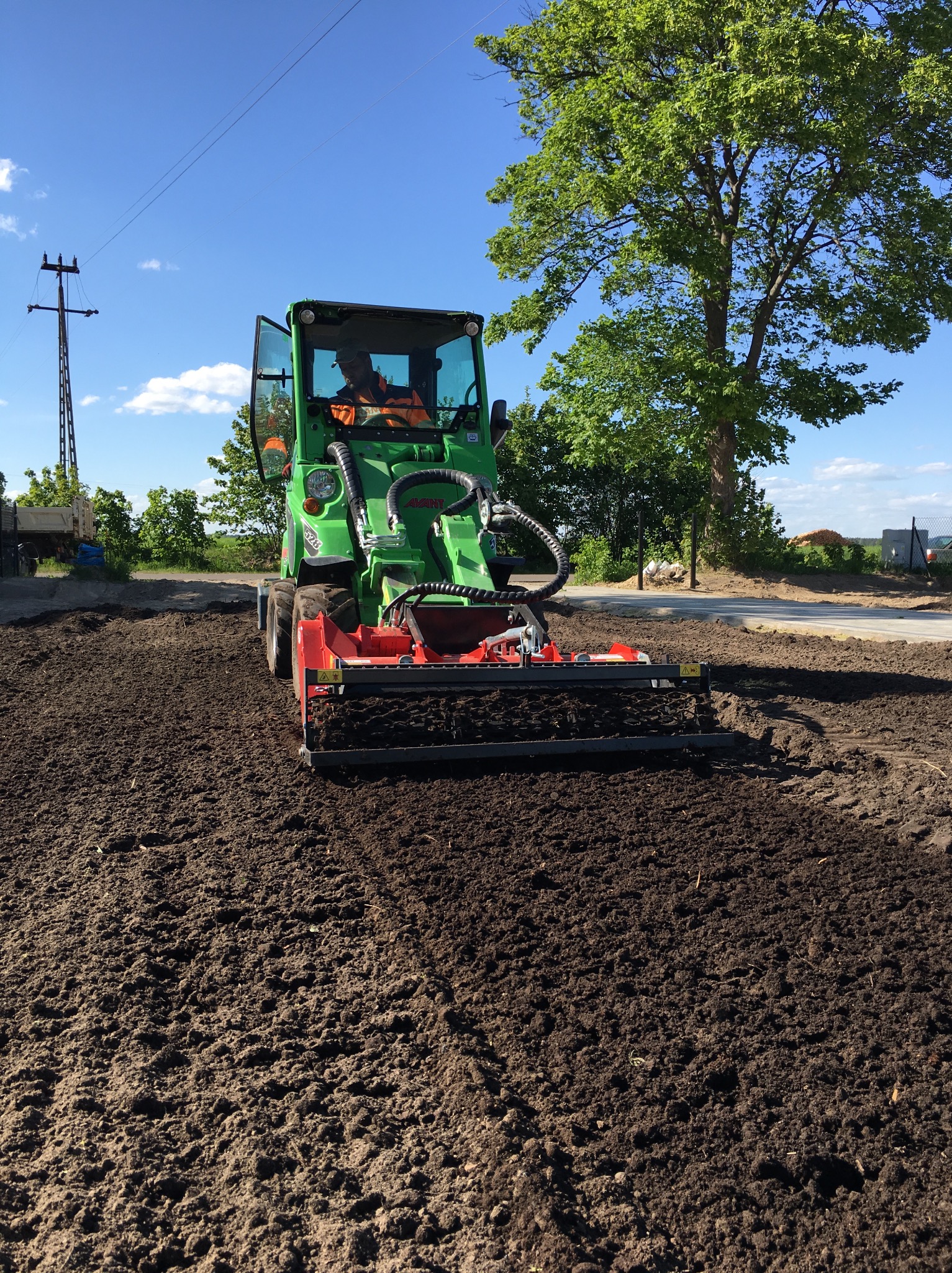 Zielony traktor z glebogryzarką spulchnia ciemną ziemię na działce. Operator w kamizelce odblaskowej. Błękitne niebo i drzewo w tle. Przygotowanie terenu pod uprawę.