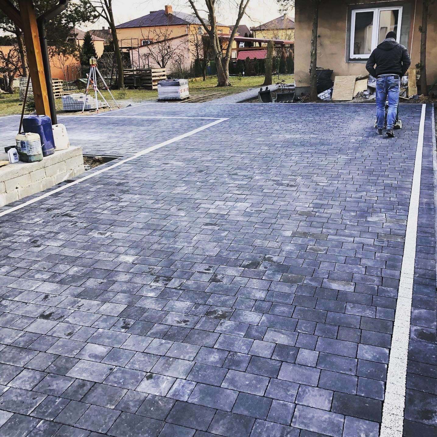 Ułożona, szara kostka brukowa z białymi liniami, narzędzia pomiarowe w tle. Mężczyzna w oddali używa maszyny do zagęszczania nawierzchni.