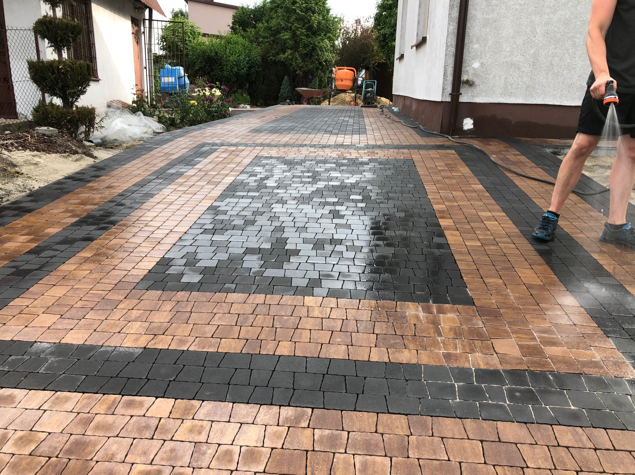 Wzorzysta nawierzchnia z kostki brukowej w odcieniach brązu i szarości, czyszczona wodą pod ciśnieniem. Widoczny fragment domu i zieleni w tle.