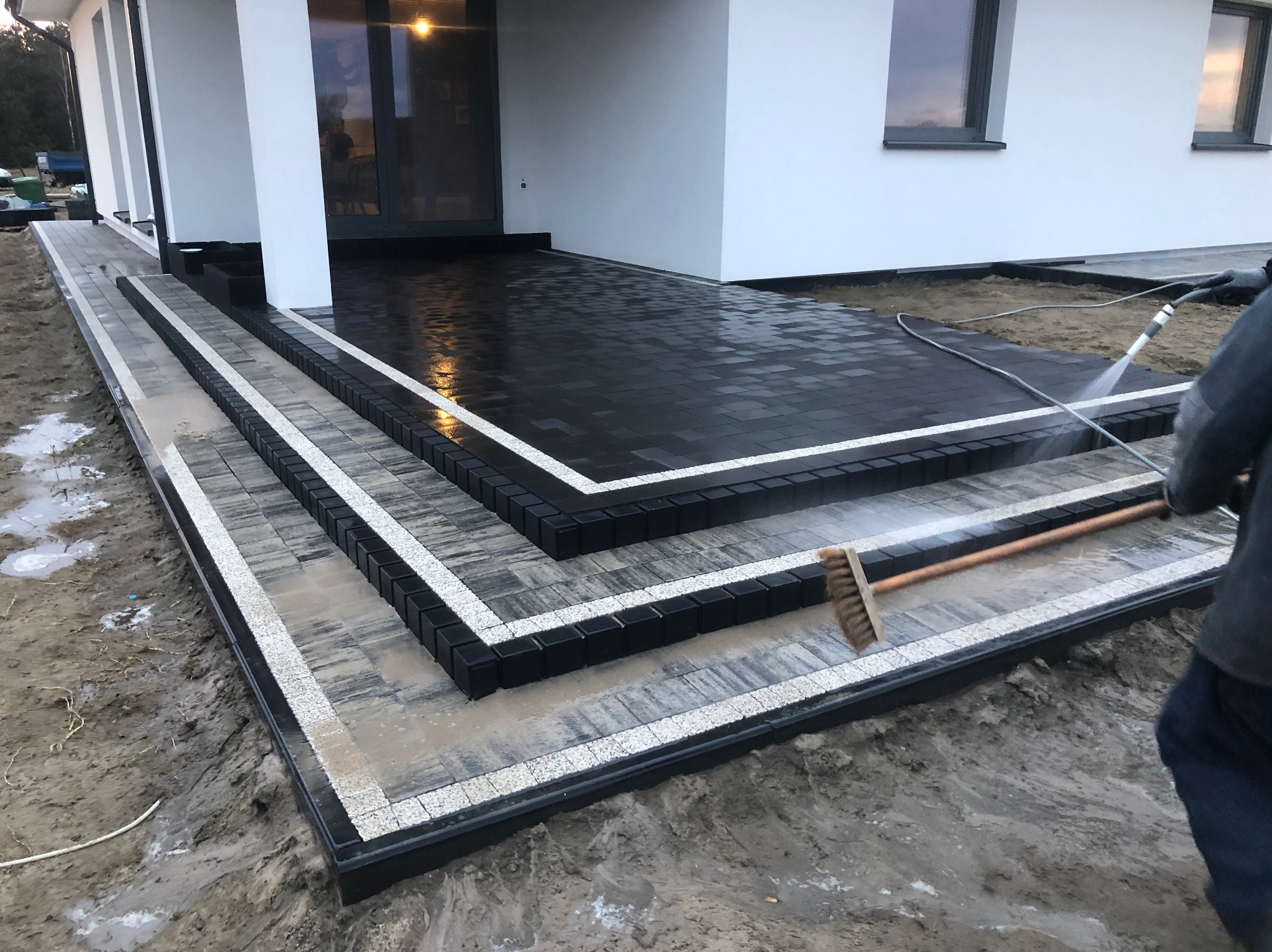 Nowoczesne schody wejściowe z kostki brukowej w odcieniach szarości i czerni, wykończone białym obramowaniem. Osoba czyści powierzchnię myjką ciśnieniową i szczotką.