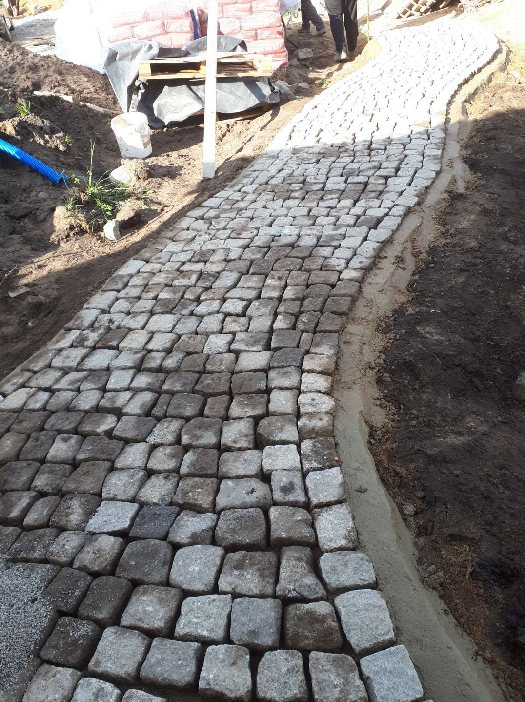 Ścieżka z kostki granitowej w trakcie układania, widoczne worki z materiałami budowlanymi w tle. Krawężnik z betonu. Częściowo ułożona nawierzchnia.