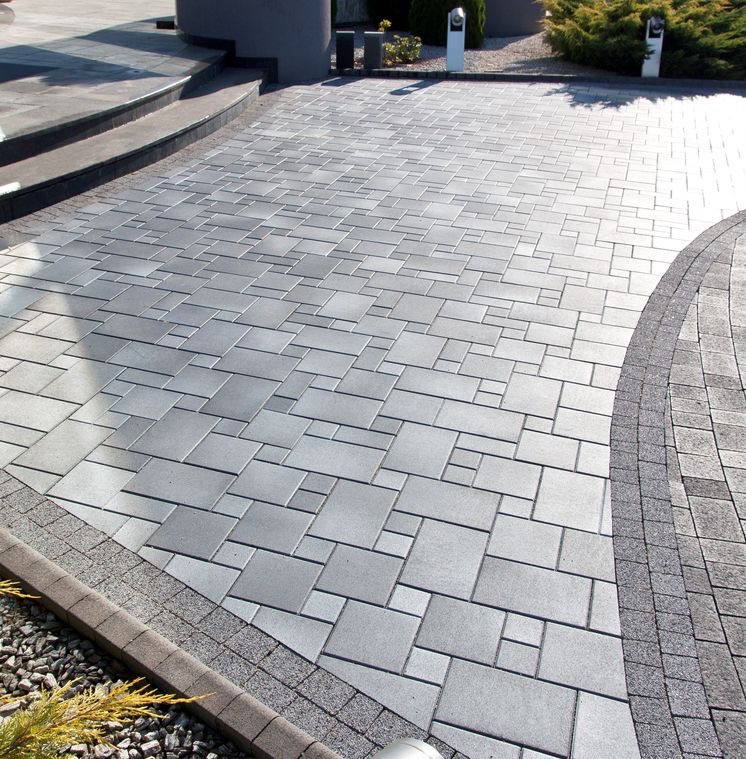 Szara kostka brukowa ułożona w geometryczny wzór, z obrzeżami z drobniejszej kostki, prowadząca do schodów i otoczona zielenią.