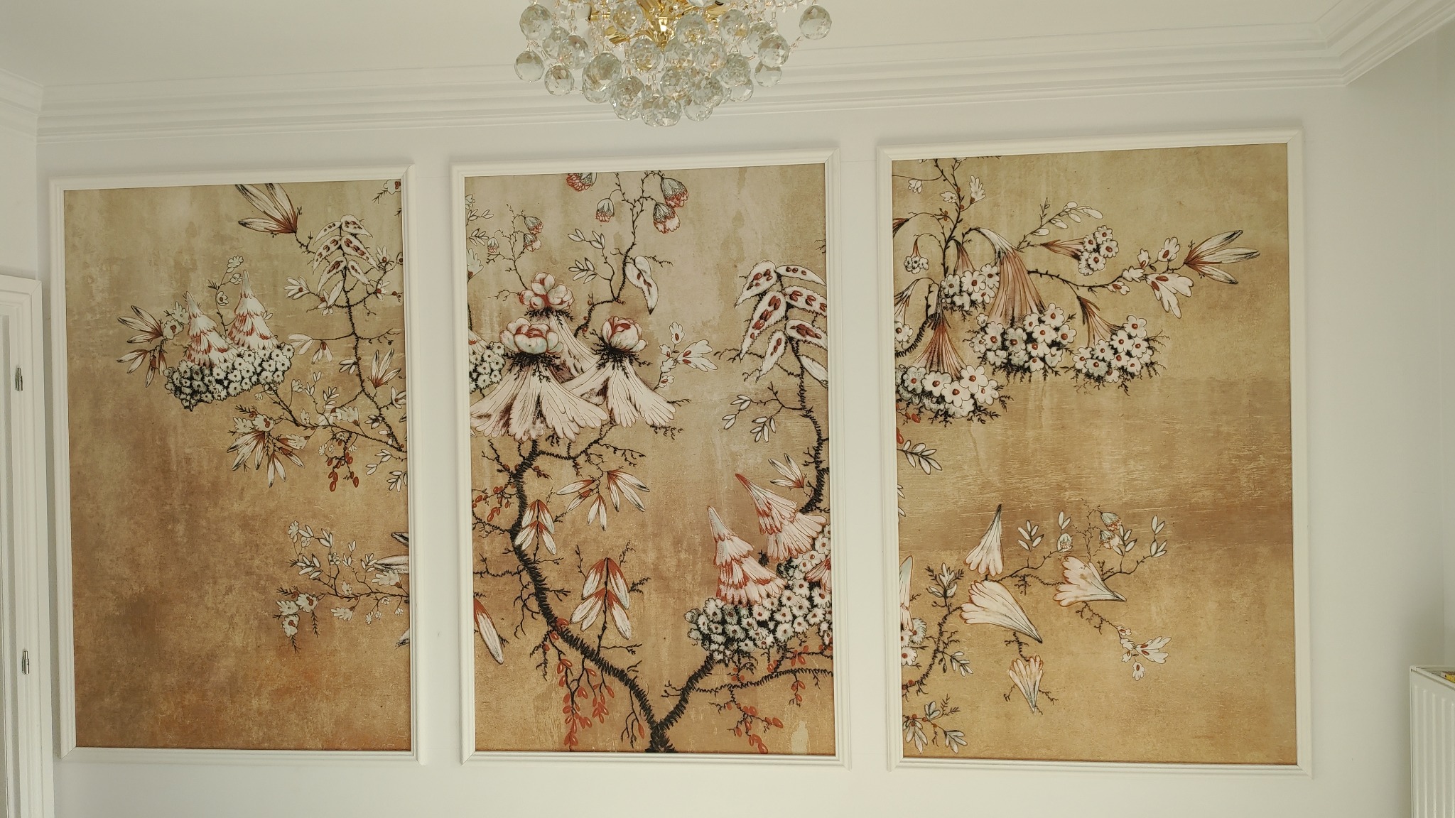 Trzy panele dekoracyjne z motywem kwiatowym w odcieniach beżu i bieli, zawieszone na białej ścianie pod kryształowym żyrandolem.