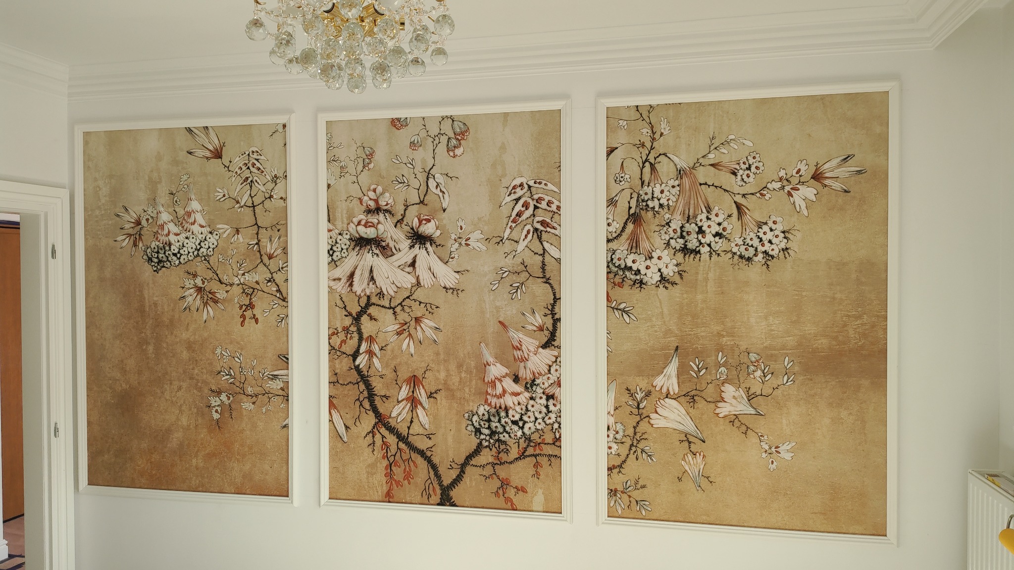 Trzy panele ścienne z motywem kwiatowym w stylu vintage, oprawione w białe ramy, oświetlone kryształowym żyrandolem, widoczne listwy przysufitowe i fragment drzwi.