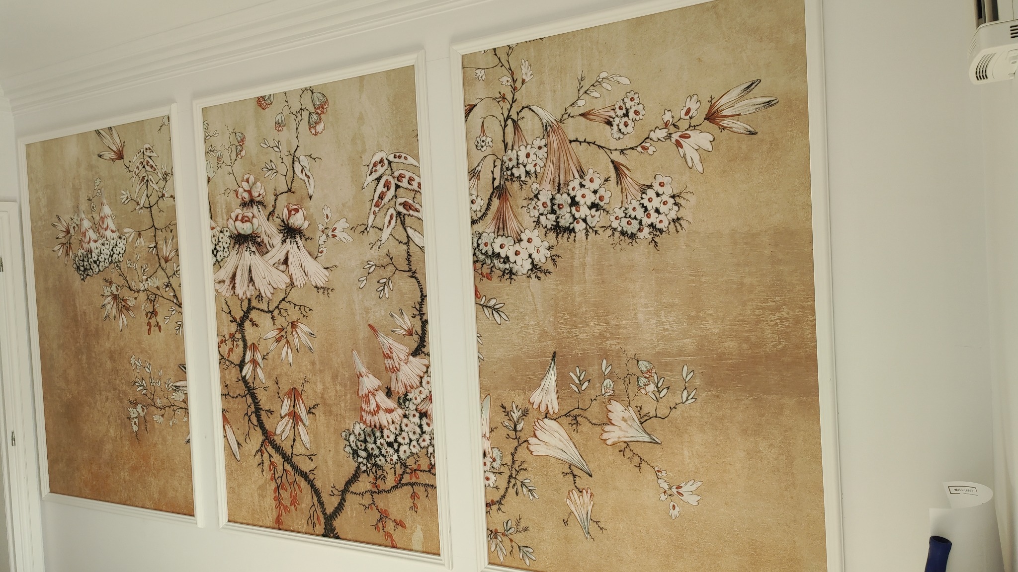 Trzy panele ścienne z motywem kwiatowym na beżowym tle, oprawione w białe ramy, umieszczone na białej ścianie z białą listwą przysufitową.