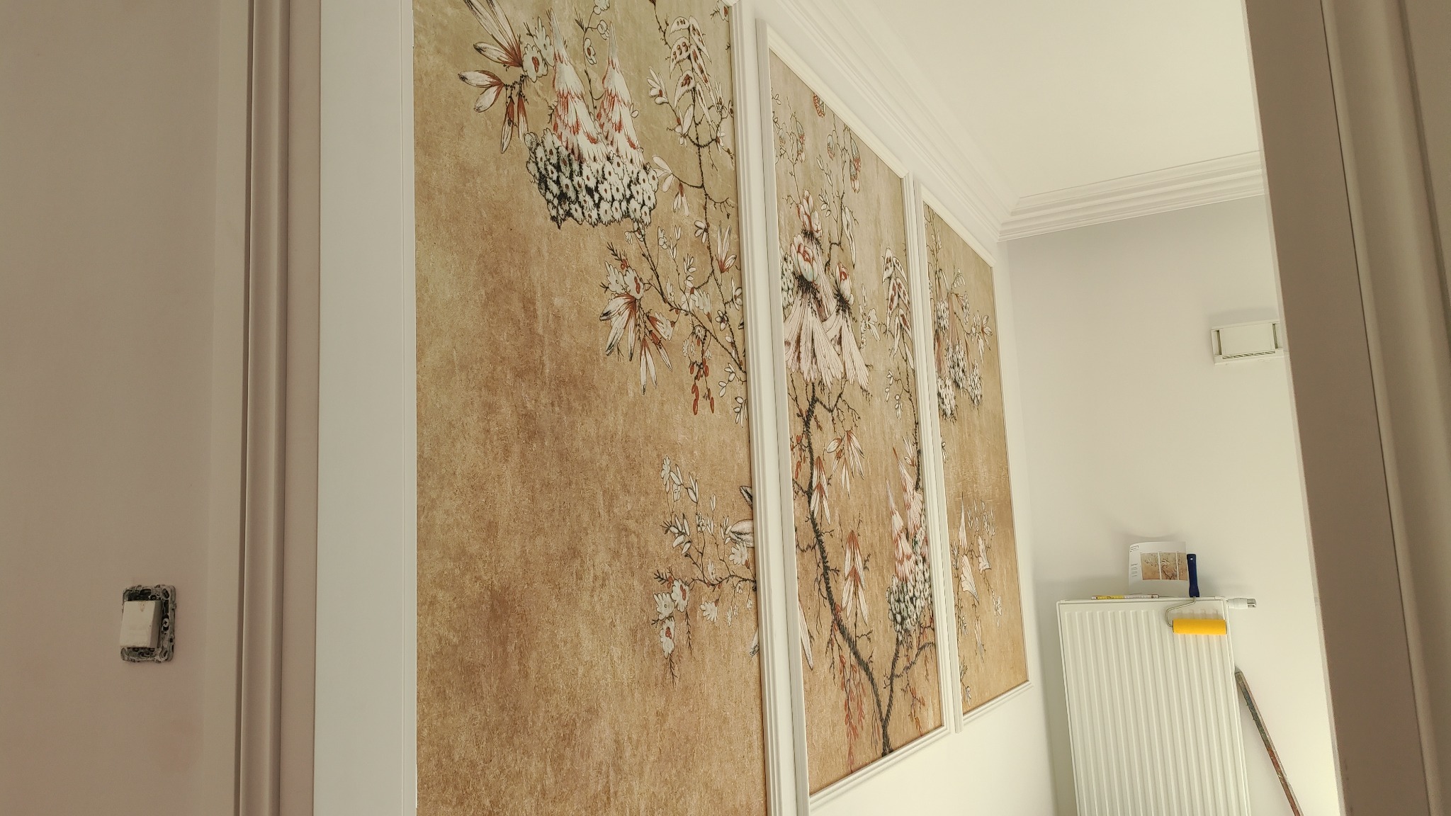 Panelowa tapeta w stylu orientalnym z motywem kwitnących gałęzi, widoczna z boku, z fragmentem grzejnika i wałka do malowania w tle.