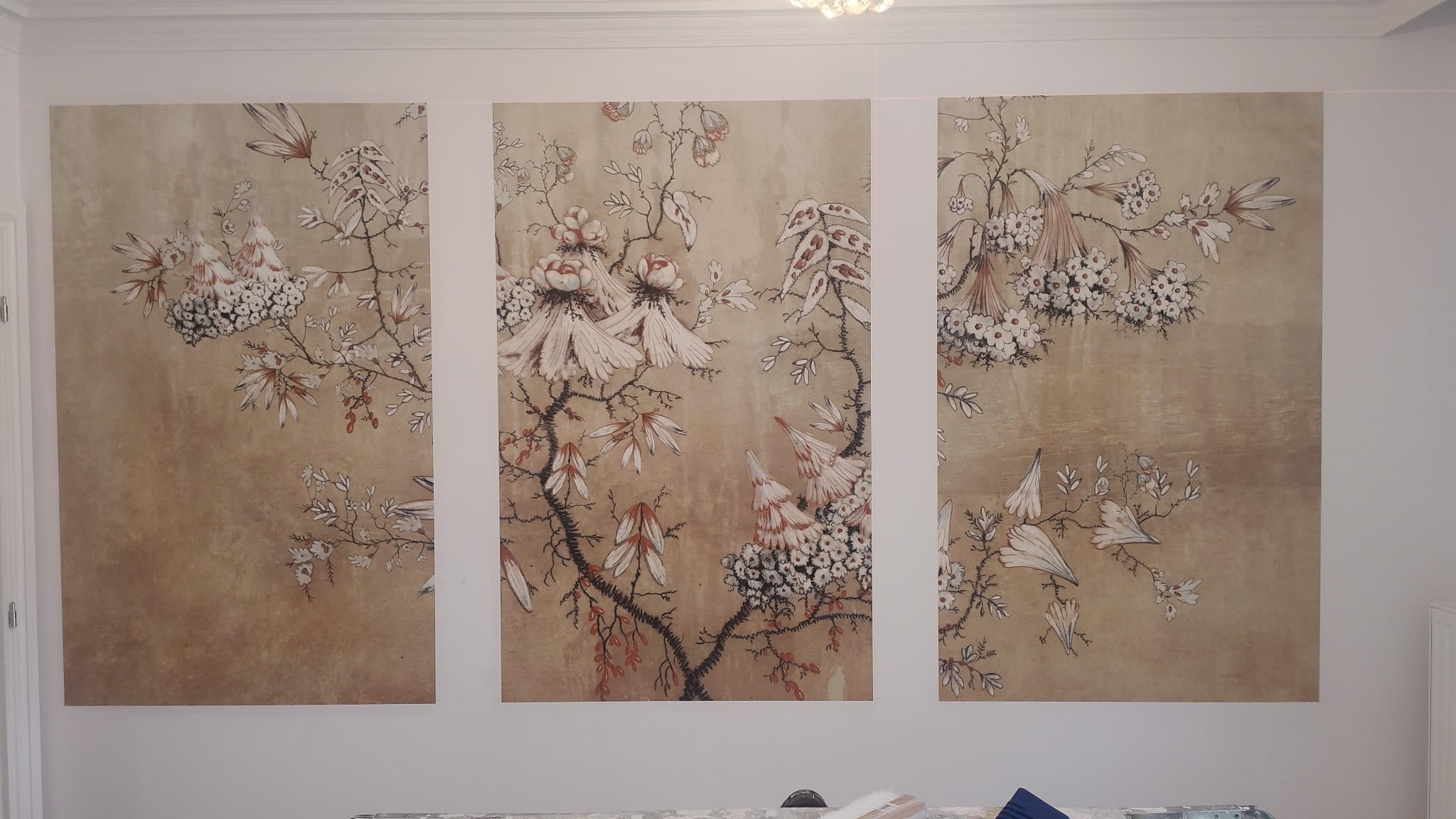 Trzy panele tapety z motywem kwiatowym na beżowym tle, tworzące spójną kompozycję na ścianie, widoczne narzędzia do tapetowania na dole kadru.