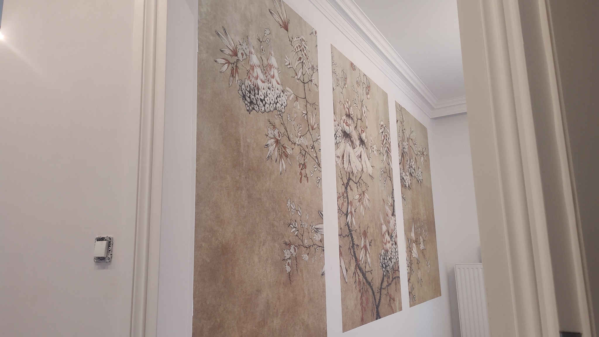 Ściana z tapetą w stylu vintage z motywem kwiatowym, podzielona na trzy panele, widoczna z perspektywy bocznej, obok biała ściana z włącznikiem światła i fragment białej futryny drzwi.