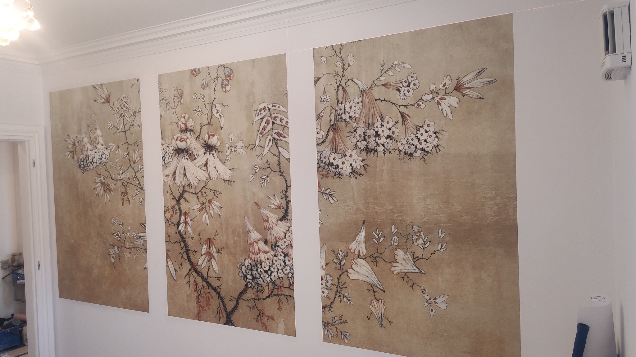 Trzy panele tapety z motywem kwitnących gałęzi na beżowym tle, zamontowane na białej ścianie w jasnym pomieszczeniu z widocznym fragmentem sufitu i klimatyzatorem.