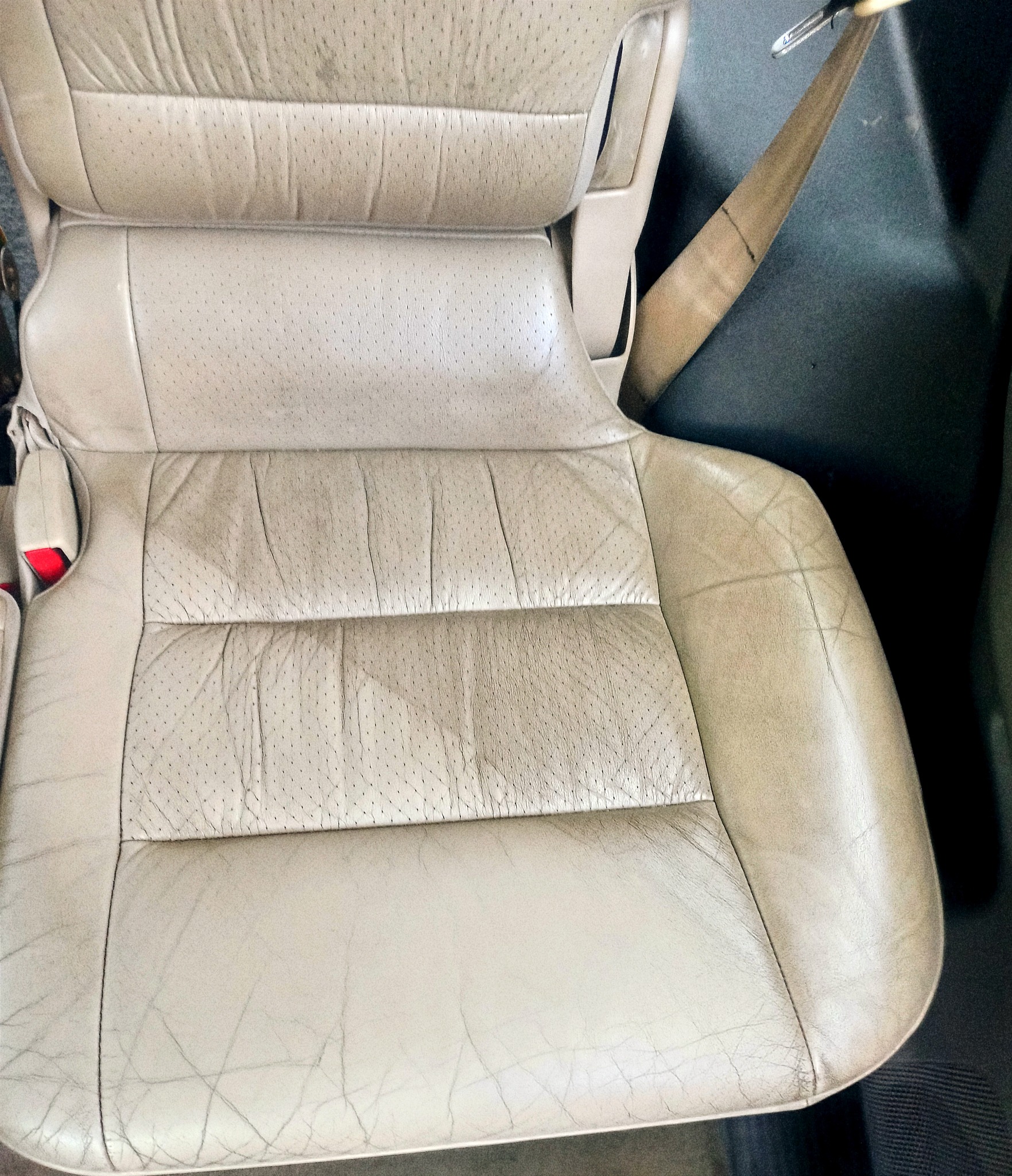 Beżowe skórzane siedzenie samochodowe z widocznymi plamami i zabrudzeniami, zapięty pas bezpieczeństwa po prawej stronie.