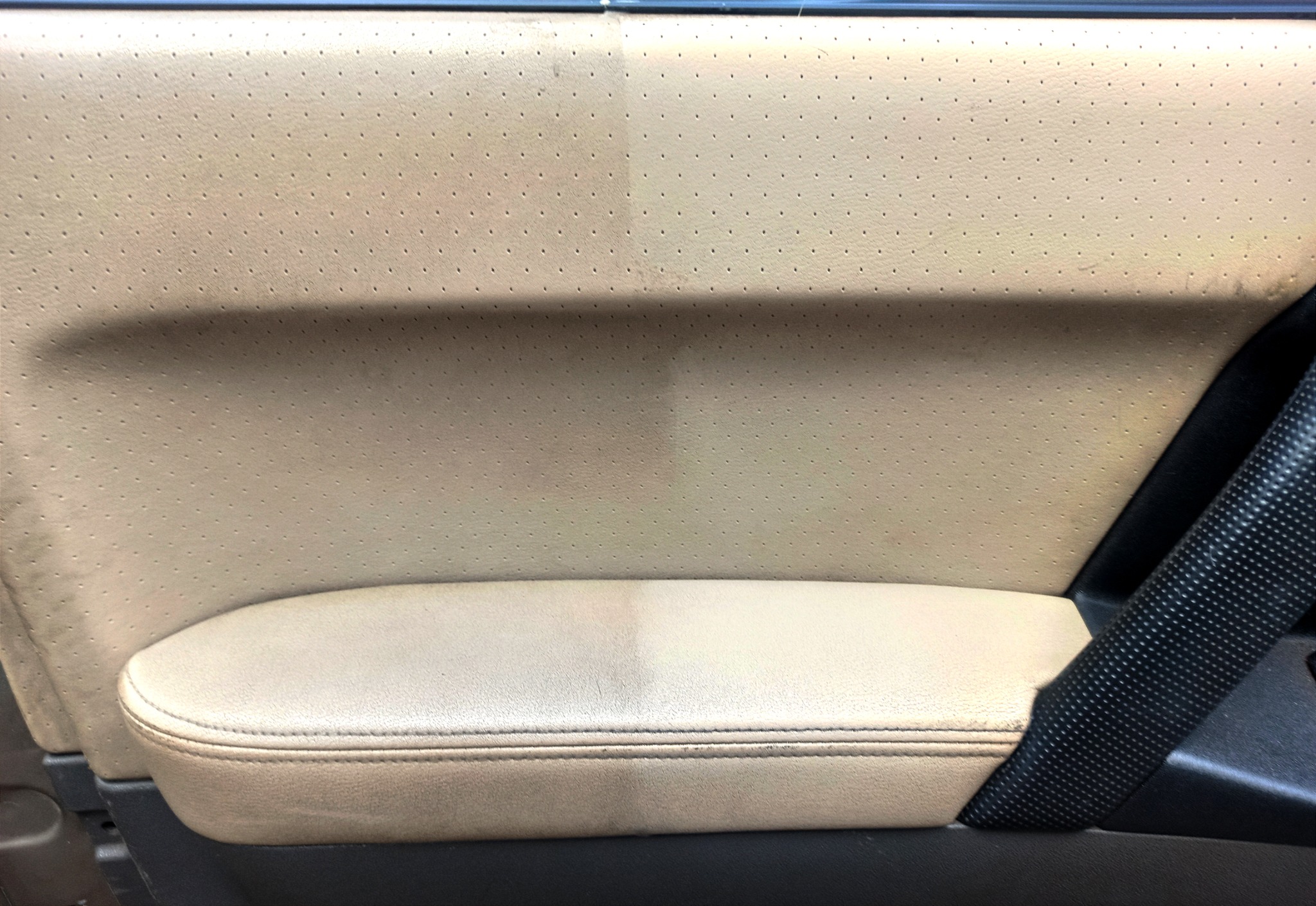 Tapicerka samochodowa w kolorze beżowym, widoczna różnica w czystości po praniu, z lewej strony zabrudzona, z prawej czysta, obok czarny pas bezpieczeństwa.