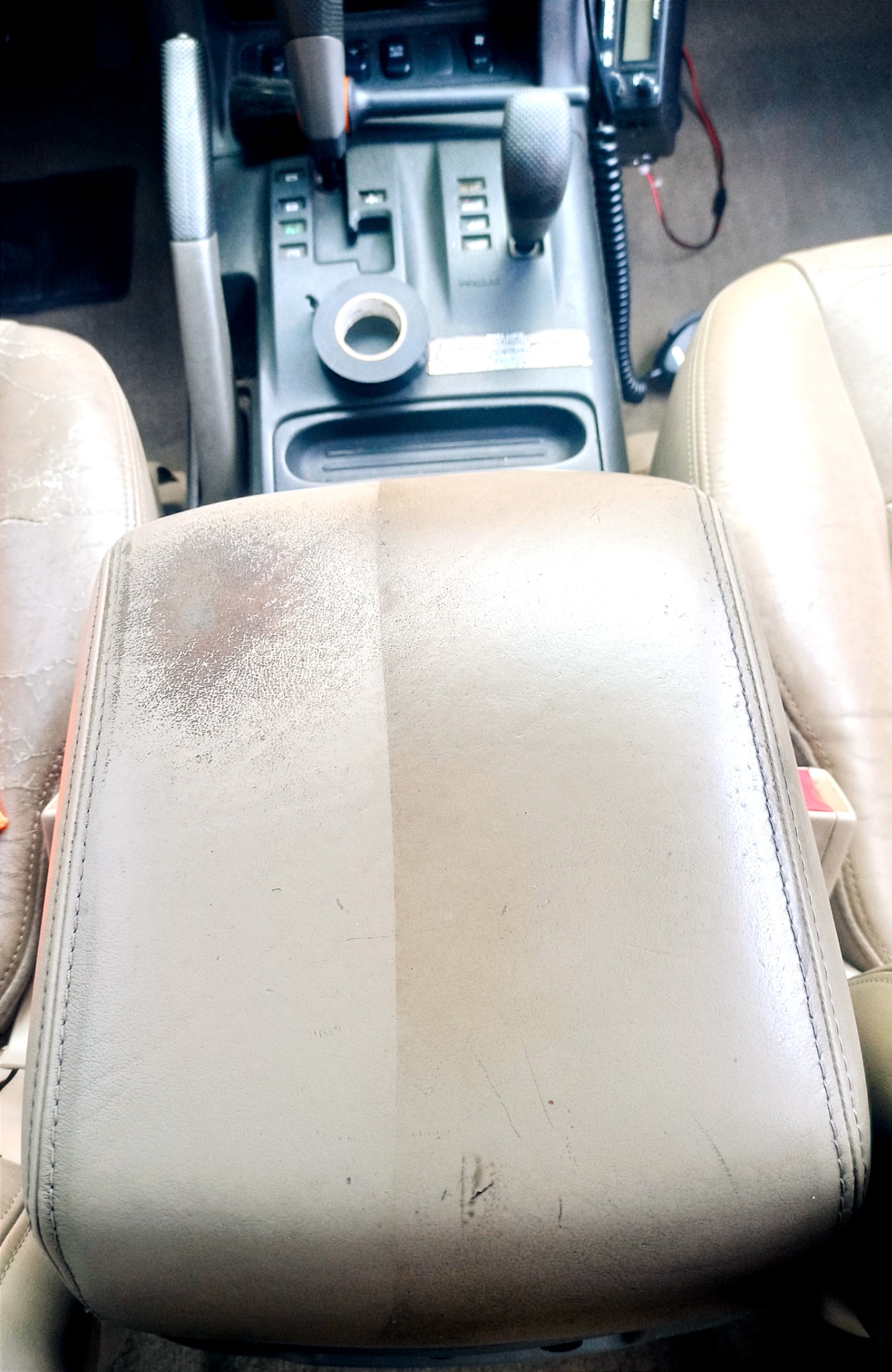 Widok z góry na beżowy podłokietnik samochodowy, ukazujący wyraźną różnicę między wyczyszczoną (prawa strona) a zabrudzoną (lewa strona) powierzchnią, z widoczną taśmą izolacyjną na konsoli środkowej.