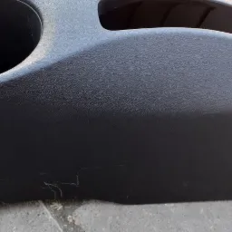 Czarny plastikowy element wnętrza samochodu z uchwytem na napoje, widoczne zarysowania i ślady użytkowania.